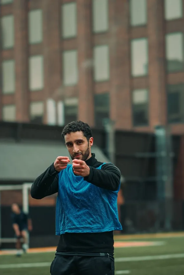 A player in a blue pinnie pointing at the camera on a turf field
