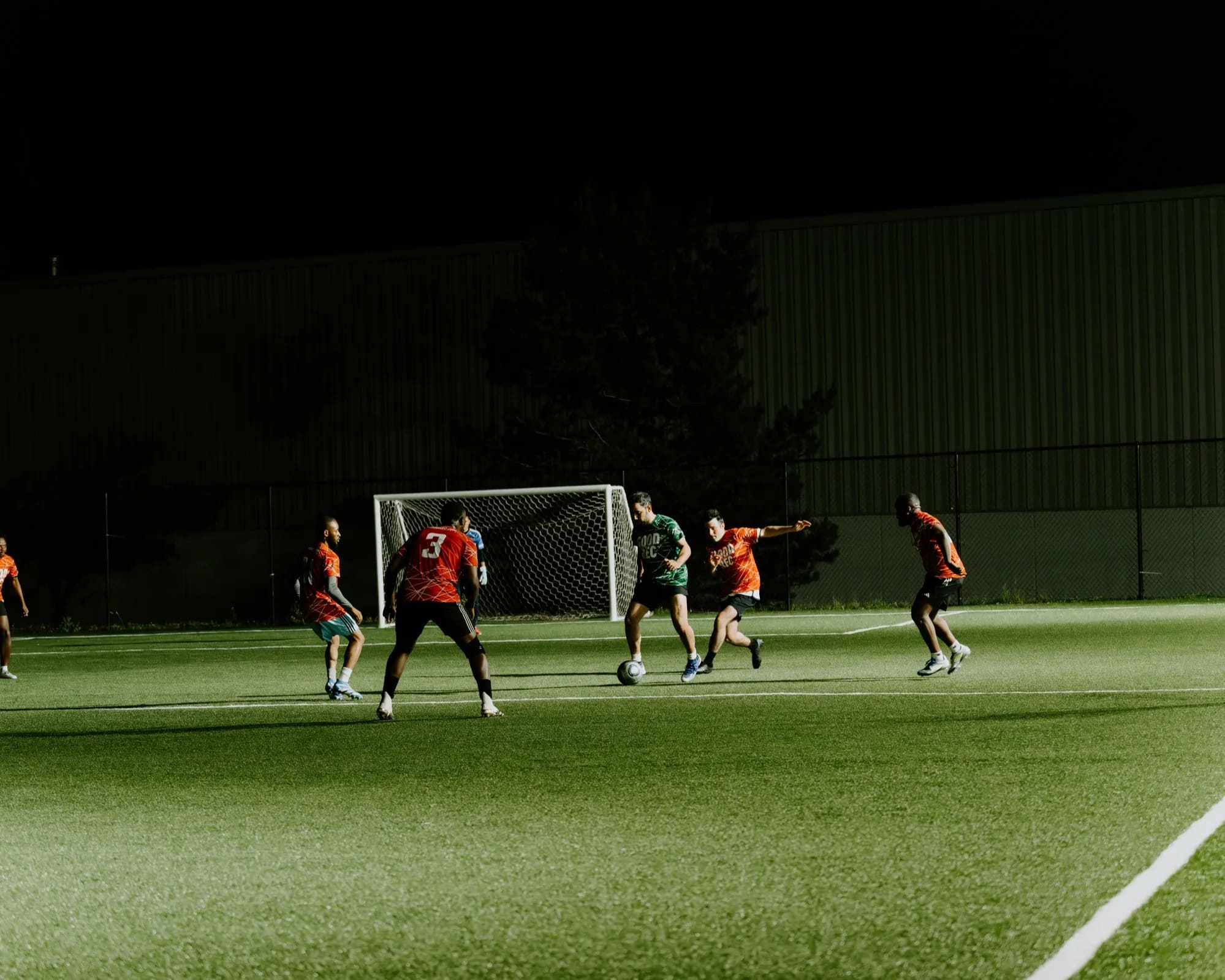 Players in GoodRec kits chasing the ball in front of goal during a floodlit evening game